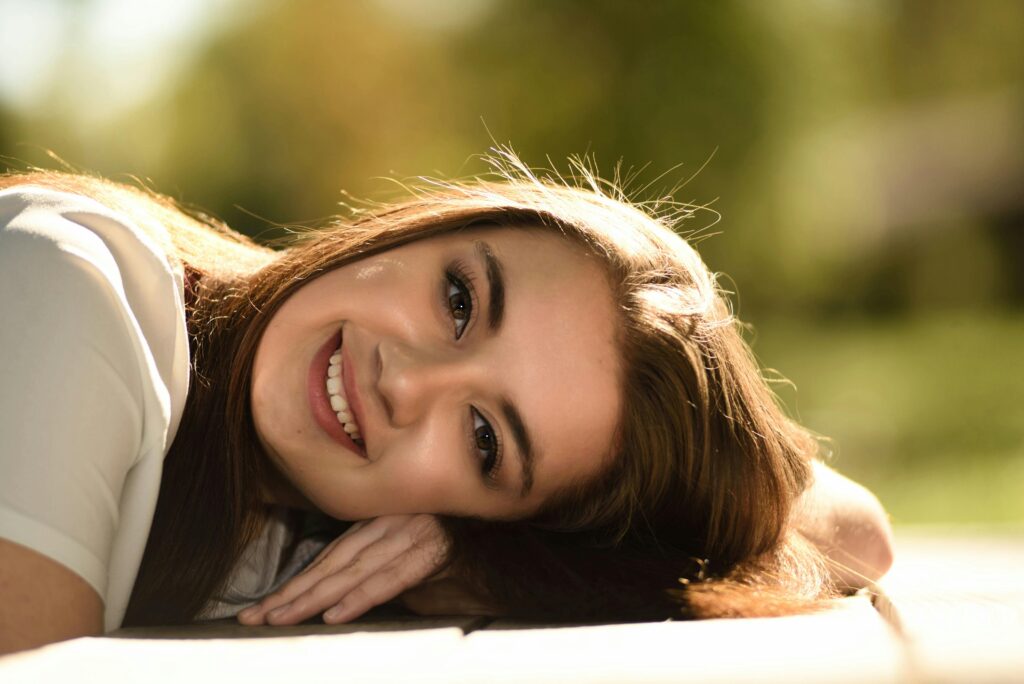 A young woman with long hair and a radiant smile lies on the grass during a sunny day.