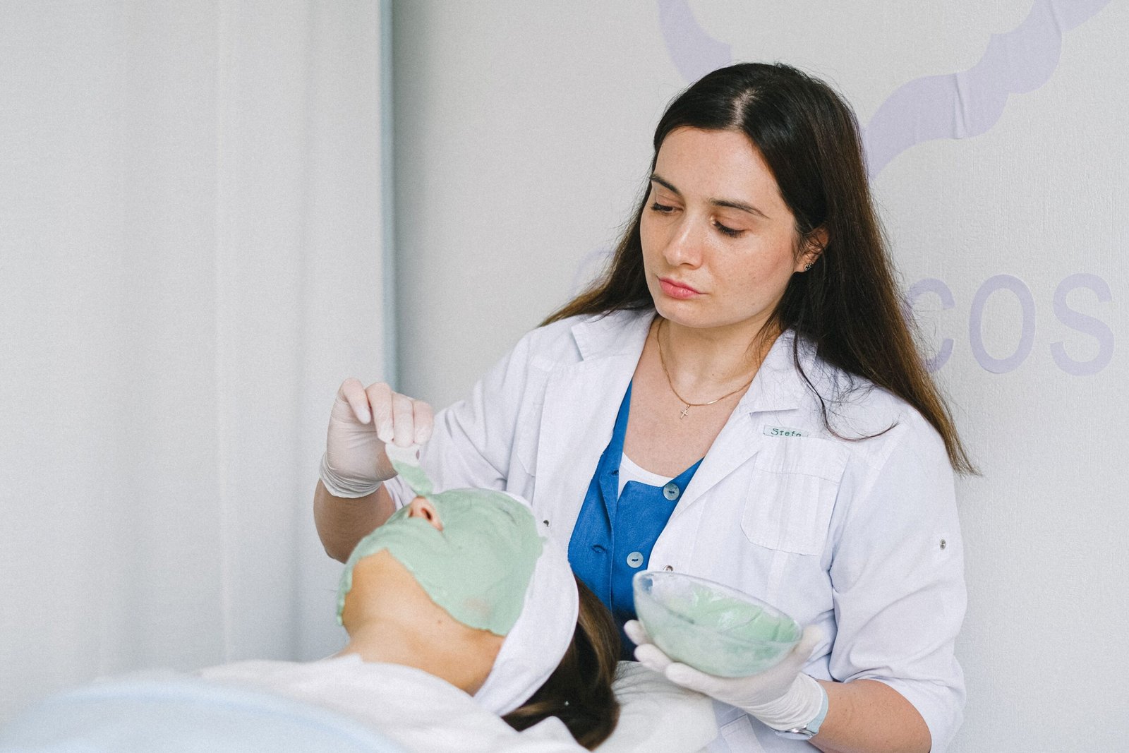 Concentrated adult female beautician applying green facial mask on woman face during spa procedure in contemporary clinic