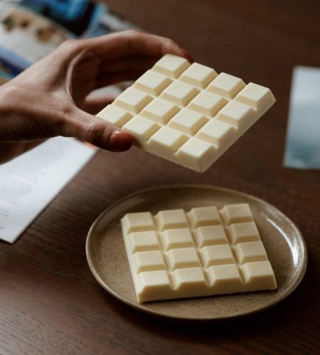 Crop faceless female holding piece of white chocolate while reading magazine and resting at home